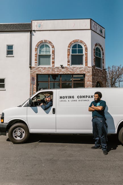 Inside and outside a white moving van with the side door open, two movers from Removal Van Harrow are visible, seated in the cab with the driver looking towards the camera and smiling. The interior of