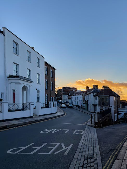 A narrow residential street in Harrow during late afternoon or early evening, showing a gentle curve with parked cars along the side. On the left side, there is a large white building with multiple windows, balconies, and a decorative entrance. The pavement features tactile paving near the curb for accessibility, and there are black metal railings along the sidewalk. The sky is partly cloudy with a warm orange glow from the setting sun, casting soft light over the scene. This environment illustrates the challenging access conditions faced during difficult removals in Harrow's narrow lanes and stairs, which Removal Van Harrow can accommodate for home relocation and furniture transport services.