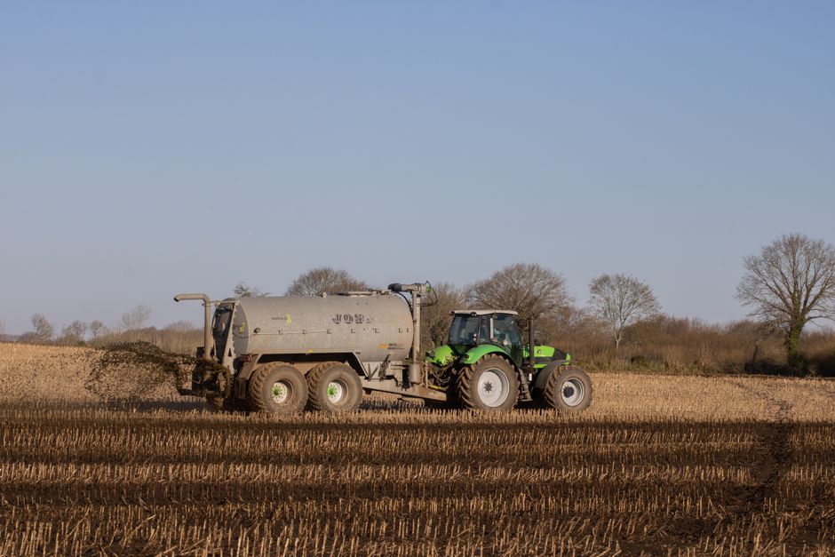 A green tractor fitted with a large, cylindrical grey water or slurry tank attached to the rear, positioned on dry, harvested farmland under clear blue skies. The tractor is actively engaged in spreading material over the stubbled field, with dark soil or fertilizer dispersing behind it. In the background, leafless trees line the horizon, indicating a late autumn or winter setting. The scene is outdoors, with natural sunlight illuminating the equipment and terrain. This image visually represents agricultural machinery used in land management, highlighting the equipment involved in operations such as soil fertilization or irrigation within farming contexts. The equipment appears robust, with large tires suitable for rough terrain, and the tank is secured with metal straps. The setting emphasizes the practical aspect of agricultural transport and operational logistics, relevant to rural or farm-based activities, but does not include furniture, moving vehicles, or indoor environments associated with house removals or home relocation.