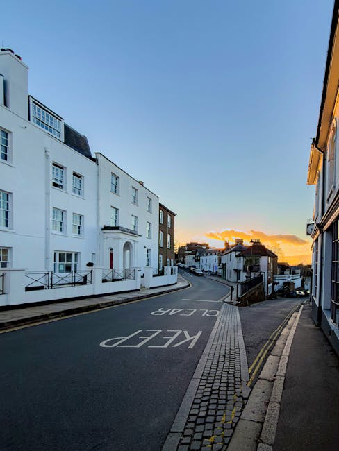 A narrow residential street in Harrow during late afternoon or early evening, showing a gentle curve with parked cars along the side. On the left side, there is a large white building with multiple windows, balconies, and a decorative entrance. The pavement features tactile paving near the curb for accessibility, and there are black metal railings along the sidewalk. The sky is partly cloudy with a warm orange glow from the setting sun, casting soft light over the scene. This environment illustrates the challenging access conditions faced during difficult removals in Harrow's narrow lanes and stairs, which Removal Van Harrow can accommodate for home relocation and furniture transport services.