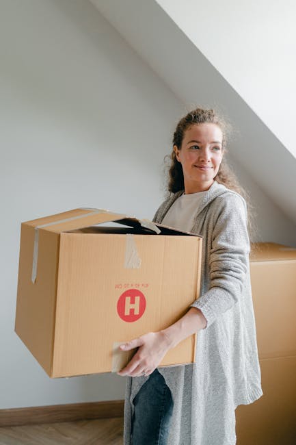 A woman with curly hair, dressed in a light grey cardigan and dark trousers, is indoors holding a large cardboard box sealed with packing tape. The box has a red 'H' logo and the words 'FRAGILE' printed on it. She is positioned near a white wall with a sloped ceiling, indicating an attic or upper floor space. Behind her, additional cardboard boxes are stacked on the wooden floor, suggesting a home relocation or packing process. The interior is brightly lit, likely from natural daylight, and the scene depicts the loading or packing stage of furniture transport or moving services, supported by the presence of packaging materials and boxes associated with house removals. Removal Van Harrow, specializing in removals and moving logistics, often manages such packing and furniture transport activities.