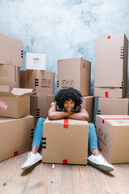 A woman with curly hair, dressed in a light grey cardigan and dark trousers, is indoors holding a large cardboard box sealed with packing tape. The box has a red 'H' logo and the words 'FRAGILE' printed on it. She is positioned near a white wall with a sloped ceiling, indicating an attic or upper floor space. Behind her, additional cardboard boxes are stacked on the wooden floor, suggesting a home relocation or packing process. The interior is brightly lit, likely from natural daylight, and the scene depicts the loading or packing stage of furniture transport or moving services, supported by the presence of packaging materials and boxes associated with house removals. Removal Van Harrow, specializing in removals and moving logistics, often manages such packing and furniture transport activities.