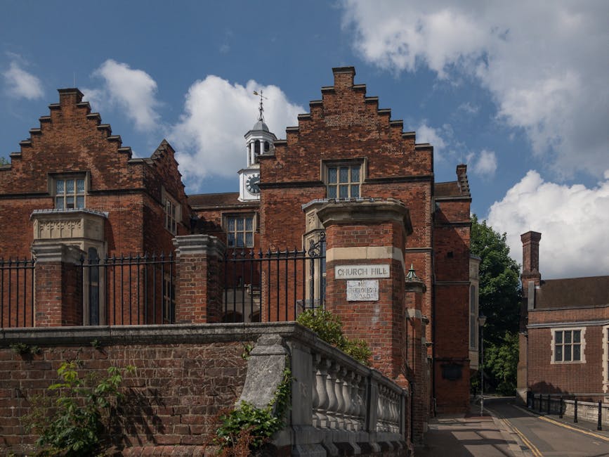 The image shows a historic red brick building with ornate gabled roofs and decorative stone window surrounds, situated behind a brick and stone wall with a black wrought iron fence on top. A white sig