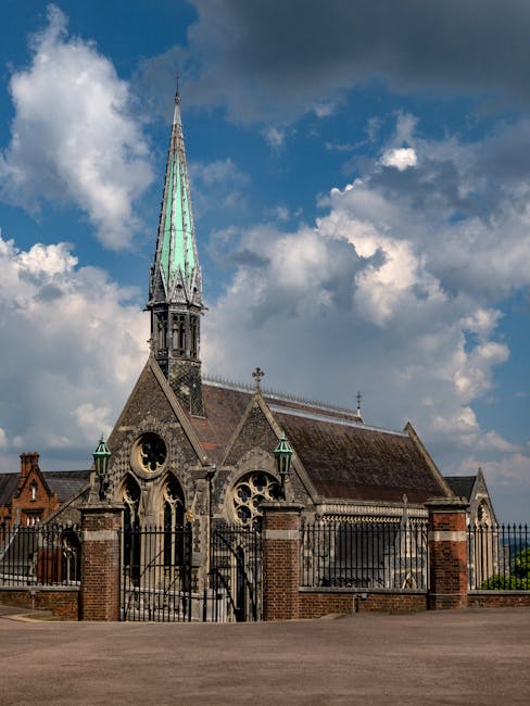 The image shows the exterior of a historic church with a pointed spire and Gothic architectural elements, including large arched windows and decorative stonework. The church is surrounded by a black metal fence supported by brick pillars, situated on a paved area. Clear weather with some clouds in the sky provides natural lighting, highlighting the texture of the stone walls and the green patina on the church's spire. The scene likely depicts a location in a residential or town area, with the church serving as a prominent feature. This setting relates to house removals and moving services, as [COMPANY_NAME] could assist with relocating items to or from historic buildings or churches, ensuring proper handling and transport during home or community relocations, including packing and loading processes involving furniture, boxes, or religious artifacts.