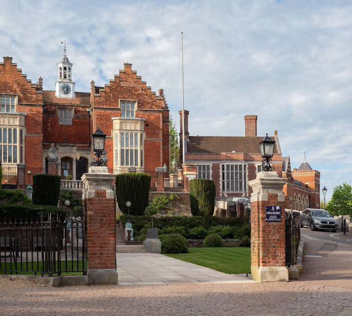 Photograph of a historic red-brick building with large windows, decorative stonework, and a clock tower, situated behind a gated driveway with brick pillars topped by traditional black iron lanterns. In the foreground, there is a paved pathway leading to the entrance, flanked by well-maintained green lawns and neatly trimmed bushes, including three tall, rounded topiary trees. The driveway curves around to the right where a grey van is parked, likely associated with house removals or furniture transport. The scene is set during daylight with a partly cloudy sky overhead. This image captures the exterior of a distinguished property, relevant to home relocation and moving services provided by Removal Van Harrow, highlighting the architectural setting typical of high-value house removals and logistical preparations for a packing and moving process.