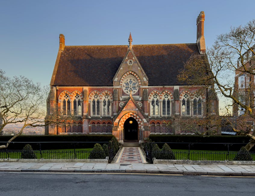 The image shows the exterior of a historic church with a pointed spire and Gothic architectural elements, including large arched windows and decorative stonework. The church is surrounded by a black metal fence supported by brick pillars, situated on a paved area. Clear weather with some clouds in the sky provides natural lighting, highlighting the texture of the stone walls and the green patina on the church's spire. The scene likely depicts a location in a residential or town area, with the church serving as a prominent feature. This setting relates to house removals and moving services, as [COMPANY_NAME] could assist with relocating items to or from historic buildings or churches, ensuring proper handling and transport during home or community relocations, including packing and loading processes involving furniture, boxes, or religious artifacts.