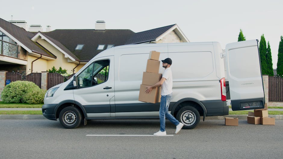 A man wearing casual clothing, including a white t-shirt, jeans, and a black cap, is carrying three cardboard boxes stacked vertically as part of the home relocation process. He is walking beside a large white removal van parked on the street in front of a suburban house with a dark roof, light-colored walls, and a wooden fence. The van's back doors are open, revealing an interior ready for furniture transport, and additional cardboard boxes are placed on the ground near the vehicle. The scene is outdoors during daylight with greenery and tall trees in the background, showcasing the loading process typical of professional removals services by Removal Van Harrow involved in packing and loading for house moves. The driver’s side window of the van is visible, and the setting emphasizes the logistical steps in furniture transport and packing during a house move. The street is quiet, and the image captures the practical aspects of a professional removals operation in Harrow.