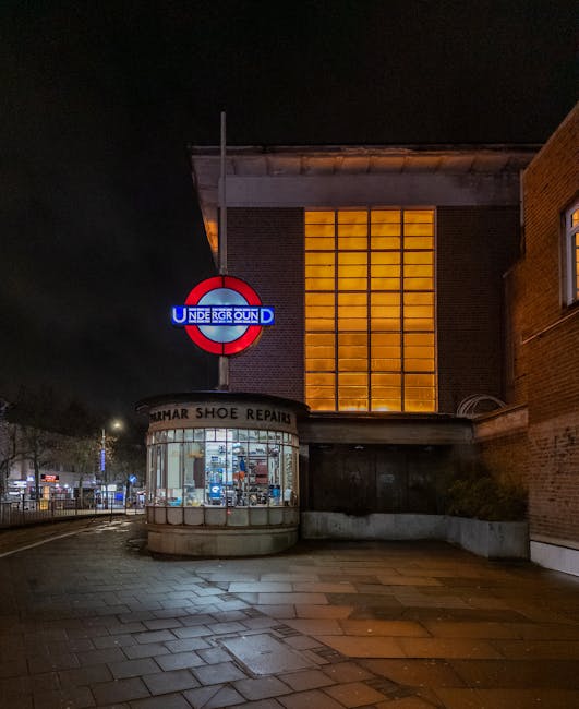 Nighttime exterior view of a brick building adjacent to a pavement illuminated by streetlights. Prominently visible is a large, illuminated red, white, and blue roundel sign with the word 'UNDERGROUND' indicating a nearby station entrance. Below the sign is a small, curved glass shop front with a sign reading 'SUMAR SHOE REPAIR', showing various shoes and repair tools inside. The pavement appears wet, reflecting some of the ambient light. The building has a large, grid-like window on the upper floor emitting a warm orange glow, contrasting with the dark sky. To the right, part of another brick building is visible. This scene could relate to an urban environment where home relocation or furniture transport services by Removal Van Harrow might operate, especially around busy transport hubs like Harrow station.